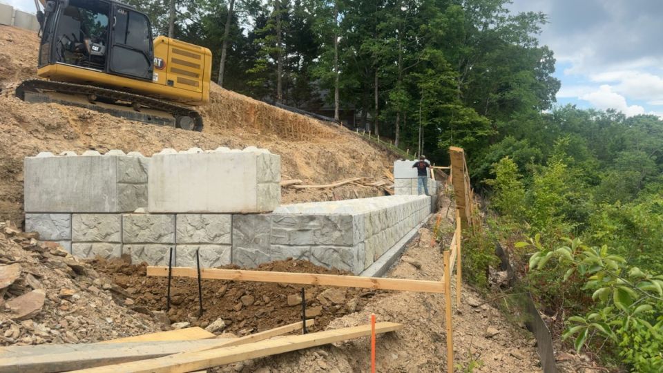 Excavator setting large stone-face concrete blocks for a retaining wall on a sloped lot