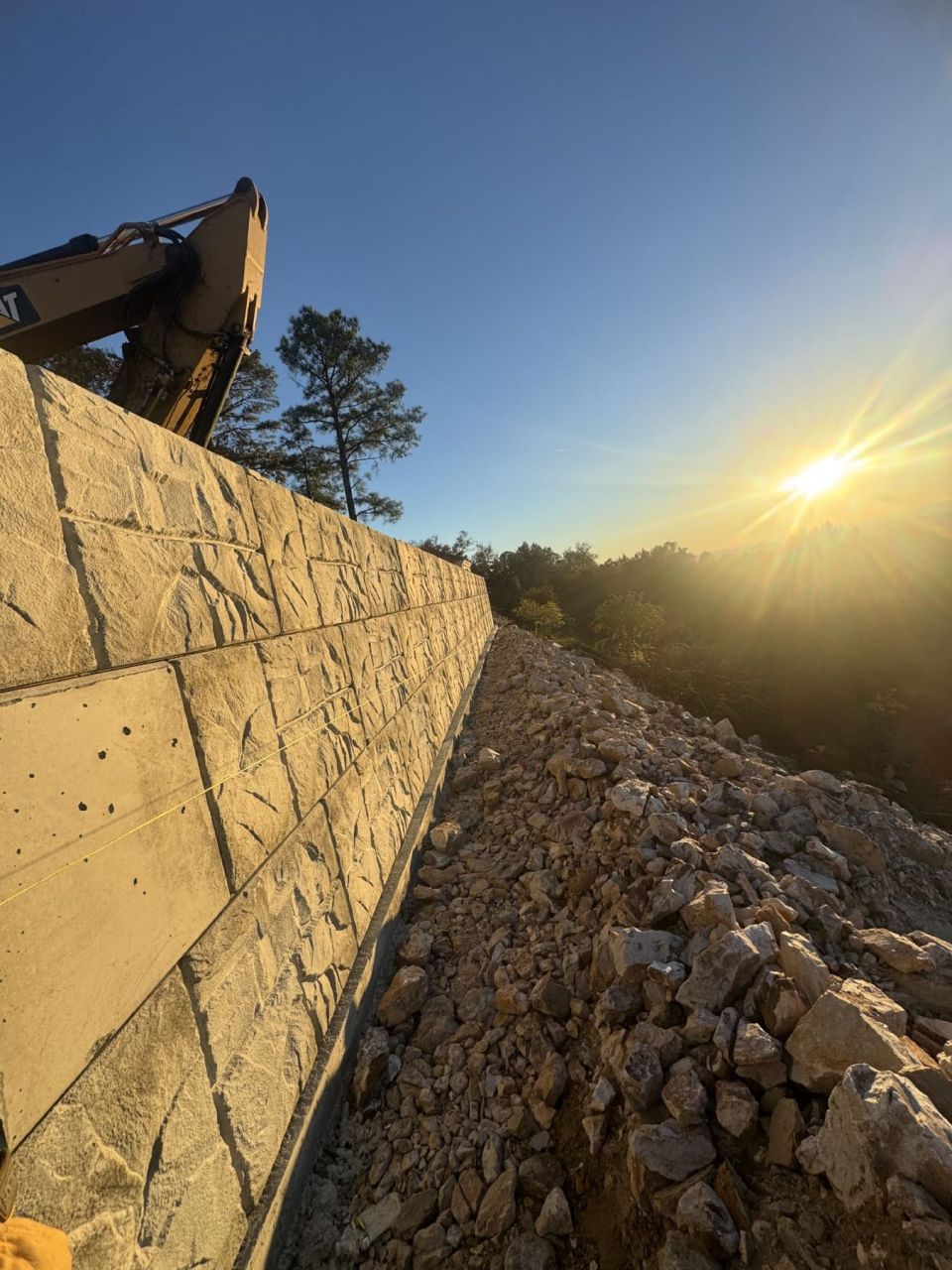 Stone-face concrete block retaining wall with rock backfill along its base and an excavator above
