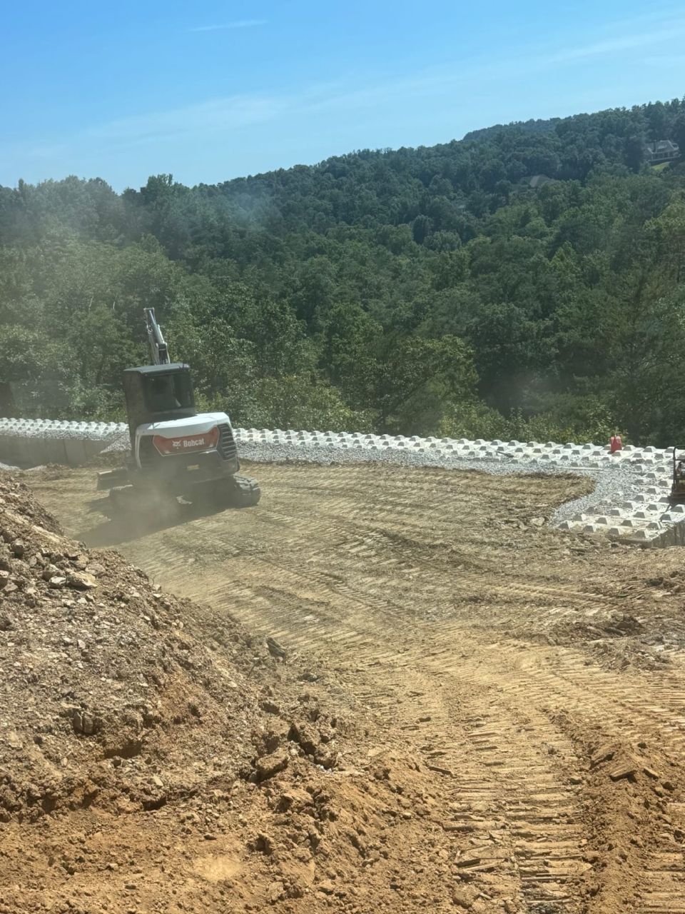 Compact excavator spreading backfill behind a curved row of new concrete retaining wall blocks