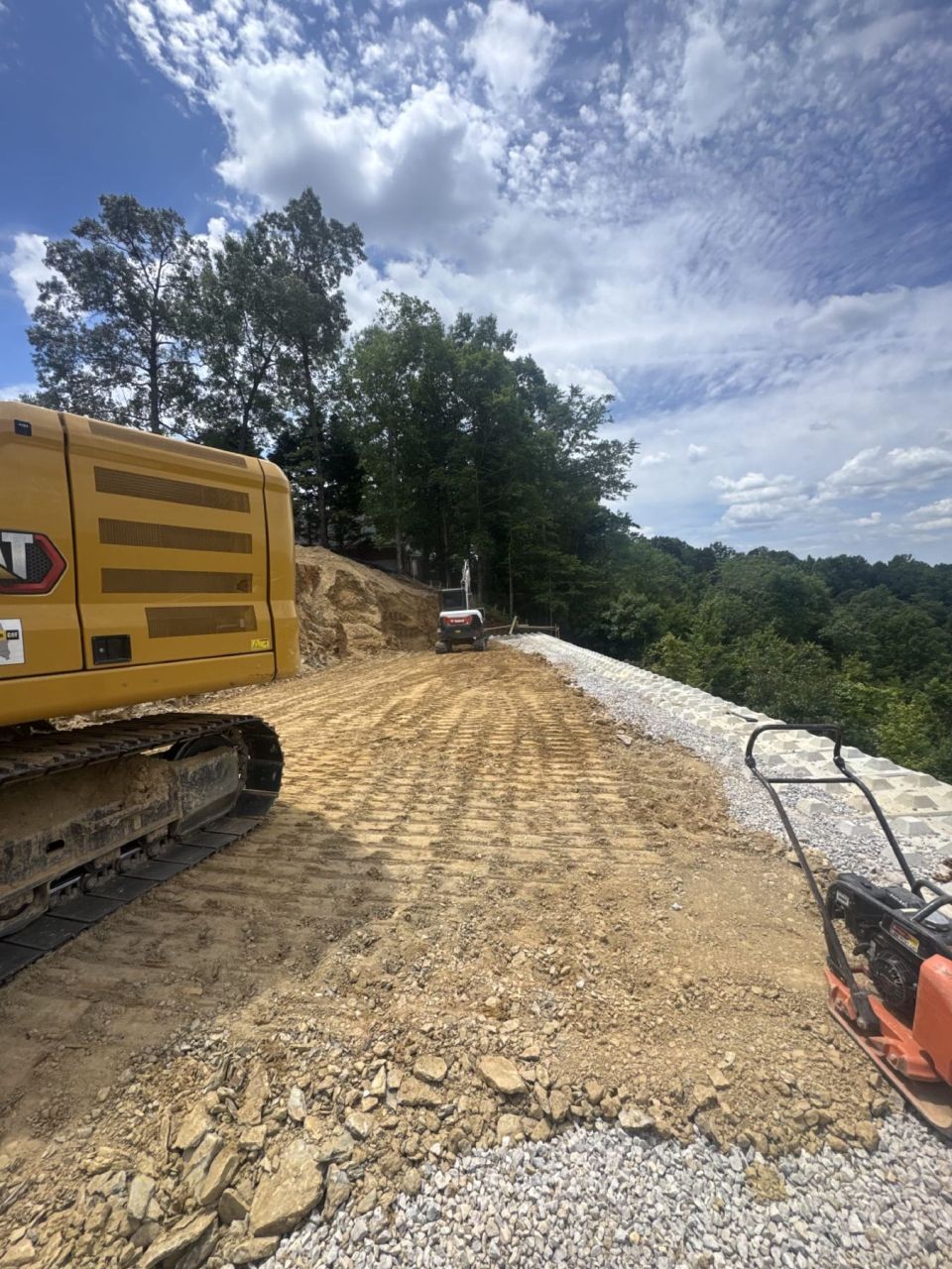 Excavator and plate compactor compacting backfill behind a retaining wall, with drainage stone along the wall